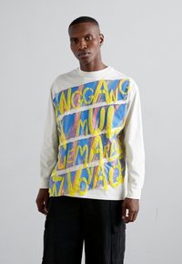 Tricou alb cu mânecă lungă, purtând un design grafic multicolor, cu text îndrăzneț galben și fundaluri abstracte albastre și roz.