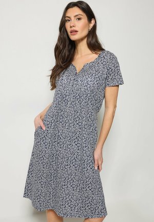 Pitkähiuksinen nainen, joka pukeutunut laivastonsiniseen polvimittaisen mekon, jossa on valkoinen kukkakuosi ja lyhyet hihat, seisoo yhdellä kädellä taskussa.