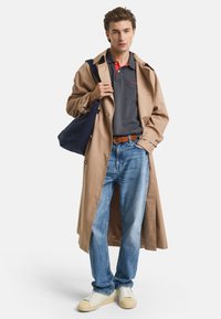 Beige Trenchcoat über einem dunkelgrauen Poloshirt mit rotem Kragen, blauen Jeans und cremefarbenen Sneakern. Trägt eine marineblaue Umhängetasche.