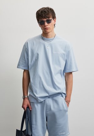 Junger Mann trägt hellblaues oversized T-Shirt und Shorts, dunkle Sonnenbrille, silbernen Schmuck, hält eine dunkle Tragetasche vor grauem Hintergrund.