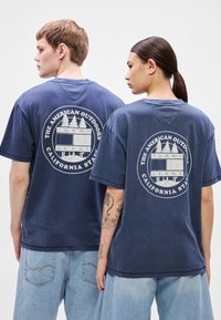 Tamsiai mėlyni medvilniniai marškinėliai su grafiniu atspaudu ant nugaros, vaizduojančiu medžius ir tekstą, derinami su šviesiai mėlynomis džinsinėmis kelnėmis. Laisvo silueto.