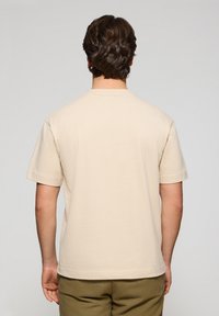 Beige bomullströja med korta ärmar och klassisk rund halsringning. Slät textur, inga synliga mönster eller dekorationer. Bakvy visas.