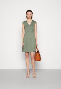 Vestido verde de manga curta com um design cruzado, corpete ajustado e saia rodada. Combinado com uma bolsa castanha e sandálias nudes de tiras.