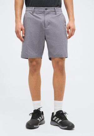 Uomo che indossa pantaloncini grigi a quadri, calzini bianchi alla caviglia, scarpe sportive nere e una maglietta nera su uno sfondo chiaro e uniforme.