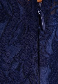 Lähikuva tummansinisestä kankaasta, jossa on monimutkaista kukka-ompelua ja yhteensopiva tummansininen vetoketjun vetotappi.