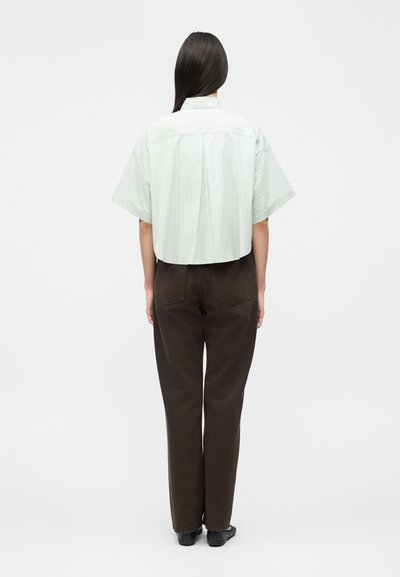 Άτομο με μακριά σκούρα μαλλιά που φοράει ανοιχτόχρωμο ριγε κοντομάνικο πουκάμισο και σκούρο καφέ παντελόνι, στέκεται με την πλάτη προς τον φακό σε απλό λευκό φόντο.