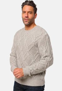 Homme avec des cheveux courts et foncés et une barbe, portant un pull en tricot léger gris et un jean noir, se tenant devant un fond blanc uni.