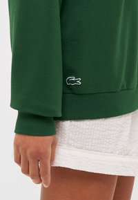 Hanorac verde cu manșete ribbate, cu un mic logo brodat al unui crocodil alb. Material neted, asociat cu pantaloni scurți texturați albi.