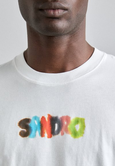 T-shirt en coton blanc avec le mot "SANDRO" en lettres floues multicolores. Design simple à col rond.