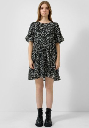 Młoda kobieta ubrana w czarną, kwiecistą sukienkę do kolan z bufkami na rękawach i czarne, wiązane botki, stojąca na jednolitym jasnym tle.