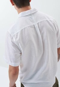 Uomo con i capelli corti e scuri che indossa una camicia di lino bianca a maniche corte, visto di spalle, con parte di un tatuaggio visibile sul braccio destro.