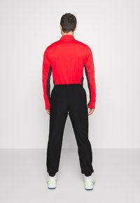 Rode sportshirt met lange mouwen en zwarte accenten, gecombineerd met zwarte elastische broek met een tailleband. De broek heeft taps toelopende pijpen en wordt gedragen met witte schoenen.