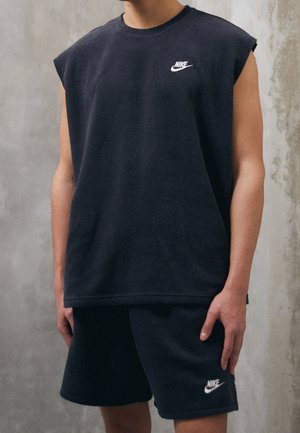 Person, die ein ärmelloses schwarzes Nike-Oberteil und passende schwarze Nike-Shorts trägt, vor einem grauen Betonwandhintergrund steht.