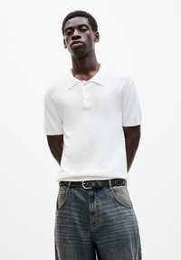 Hvid kortærmet polo med klassisk krave og tre-knap lukning, parret med blå denimjeans med en falmet vask og sort bælte.