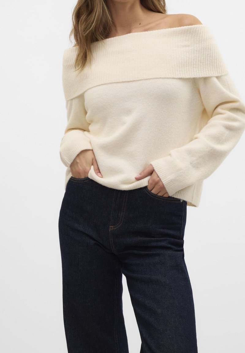 Femme portant un pull crème à épaules dénudées avec manches longues et un jean taille haute foncé, mains dans les poches, sur fond uni.