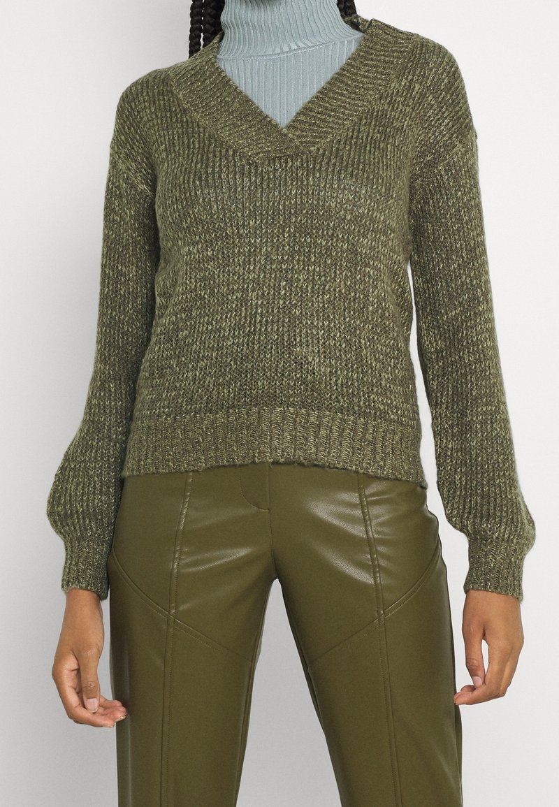 Maglione lavorato a maglia verde oliva con scollo a V, polsini a costine e tessuto testurizzato, abbinato a pantaloni aderenti lucidi verde oliva.
