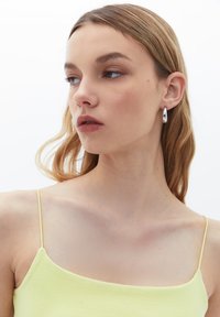 Camisole gialla a costine con spalline sottili, abbinato a orecchini a cerchio in argento. Il modello ha lunghi capelli castano chiaro e un trucco neutro. Sfondo neutro.