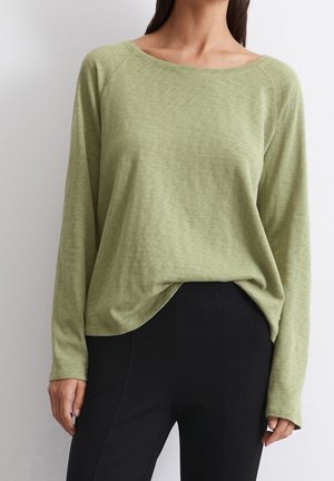 Haut à manches longues de couleur vert clair, confectionné dans un tissu doux avec une coupe légèrement ample. Dispose d'un large col bateau et d'un ourlet courbé. Pantalon noir visible.