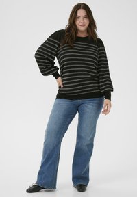Mujer con un suéter de mangas abullonadas negro y gris a rayas, jeans azules y bailarinas negras, de pie con la mano en la cadera.