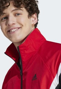 Giacca atletica rossa con colletto alto, chiusura con zip e accenti in bianco e nero. Il materiale sembra lucido con una finitura testurizzata.