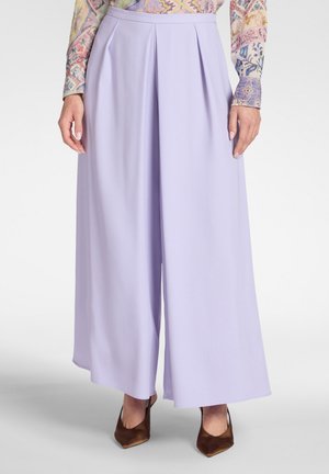 Mujer con pantalones lila de talle alto y pierna ancha, con pliegues delanteros, y tacones marrones de punta afilada, combinados con una blusa de manga larga con estampado.