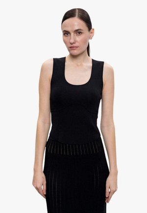 Donna con capelli castani lisci raccolti indietro, indossa un top nero senza maniche e una gonna nera coordinata con motivi verticali, in piedi davanti a uno sfondo bianco.