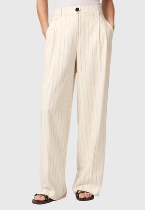 Kremowe spodnie o szerokich nogawkach z cienkimi pionowymi prążkami, z przodu zapięte na guziki i zamek błyskawiczny, noszone z brązowymi sandałami na otwartym palcu, ręce w kieszeniach.