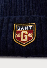 Navy knit beanie with ribbed texture, featuring a shield patch in red, yellow, and white. The patch reads "GANT 1949" with a central "G."