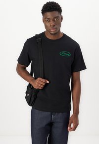 Svart bomulls T-skjorte med grønt logo på brystet, kombinert med mørke denimbukser og en liten svart crossbody-veske.