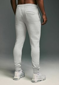 Grijze joggingsbroek met een taps toelopend ontwerp, een elastische tailleband en een zijzak, gecombineerd met witte sportschoenen op een neutrale achtergrond.