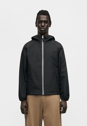 Jeune homme debout, portant une veste à capuche noire zippée et un pantalon ample couleur sable sur un fond clair uni.