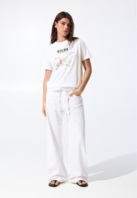 T-shirt blanc avec le texte "GILDA" en vert et un graphique de poivrons, associé à un pantalon blanc ample et des sandales noires. Tissu lisse, coupe décontractée.