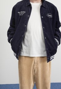 Giacca di velluto a coste blu navy con polsini a strisce bianche, caratterizzata da testo ricamato e logo, indossata sopra una t-shirt bianca e un pantalone di velluto a coste beige.