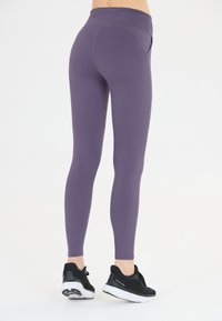 Kvinna med lila tajta leggings och svarta träningsskor, stående mot en plain vit bakgrund, visad bakifrån.