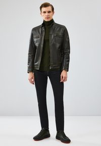 Veste en cuir noire avec deux poches poitrine, col roulé vert, jeans foncés et baskets noires avec des accents orange. Texture lisse et coupe ajustée.