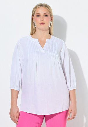 Blonde Frau trägt eine weiße Langarmbluse mit Biesen-Details und pinke Hose, steht an einer weißen Wand.