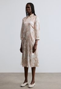 Φόρεμα ανοιχτό ροζ με λουλουδάτο μοτίβο, γιακά και μανίκια τριών τετάρτων, από λείο ύφασμα. Φορεμένο με μπεζ παπούτσια τύπου slip-on.