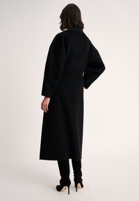 Długi czarny płaszcz z wyraźnie uformowanym kołnierzem i oversize'owymi rękawami, wykonany z mieszanki wełny, charakteryzujący się czystą sylwetką i gładką fakturą.