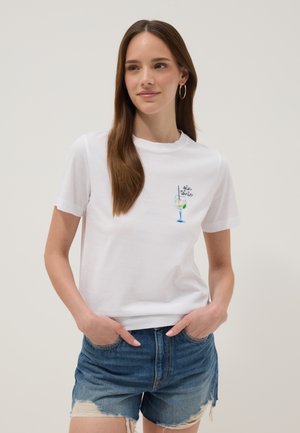 Young woman with long brown hair, wearing gold hoop earrings, a white t-shirt with a gin tonic illustration, and distressed denim shorts.