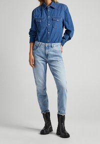 Tummansininen farkkupaita, jossa on kaksi rintataskua, yhdistettynä vaaleansinisiin farkkuihin ja mustiin nilkkabootseihin, joissa on paksu pohja. Teksturoitu kangas.