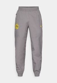 Jogging gris avec une taille élastique, des poches zippées et un détail de logo jaune avec des étoiles sur la jambe gauche et le logo de la marque sur la jambe droite.