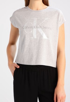 Kobieta nosząca jasnoszary krótki t-shirt Calvin Klein Jeans i czarne spodnie, z jedną ręką w kieszeni.
