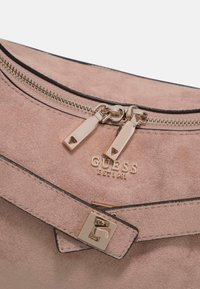 Gros plan sur un sac Guess en daim rose avec double curseur de fermeture éclair doré, texte du logo et bandoulière décorative avec boucle carrée.