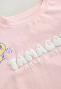 Lyserød t-shirt med korte ærmer, der har et gult karaktergrafik og hvid, rund tekst, som står "TAMAGOTCHI HAVE A HAPPY."
