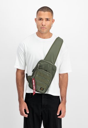 Olive green sling bag with a rectangular shape, padded texture, a front zip pocket, and a decorative red strap with text.