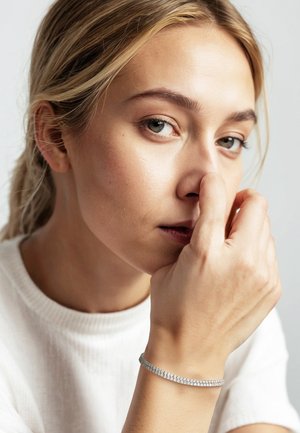 Silbernes Armband mit mehreren kleinen, spiegelnden Steinen, das an einer Hand getragen wird, die nahe am Gesicht ruht, vor einem hellen Hintergrund.