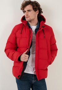 Rød puffjakke med hætte, prydet med et quiltet design, forlommer og sorte detaljer. Båret med lynlåsen åben over en hvid skjorte og denim.