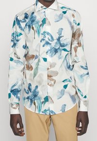 Chemise blanche boutonnée avec un motif floral aquarelle en bleu, vert et marron. Manches longues, col classique et coupe ajustée.