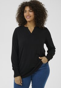T-shirt noir à manches longues avec un décolleté en V, présentant des détails froncés aux poignets. Tissu doux, coupe décontractée, associé à un jean en denim bleu.