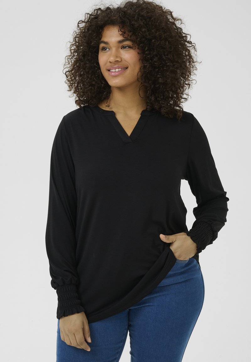 T-shirt noir à manches longues avec un décolleté en V, présentant des détails froncés aux poignets. Tissu doux, coupe décontractée, associé à un jean en denim bleu.